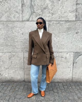 Woman wears jeans, a turtleneck, 80's style blazer and tan loafers.