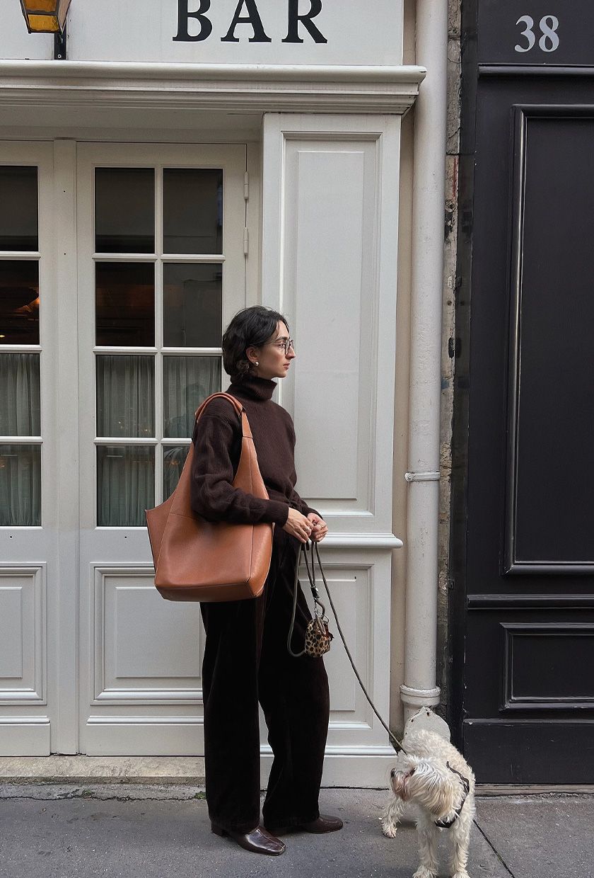A woman wearing a chocolate brown turtleneck with brown velvet trousers, brown ballet flats, and a light brown tote bag.