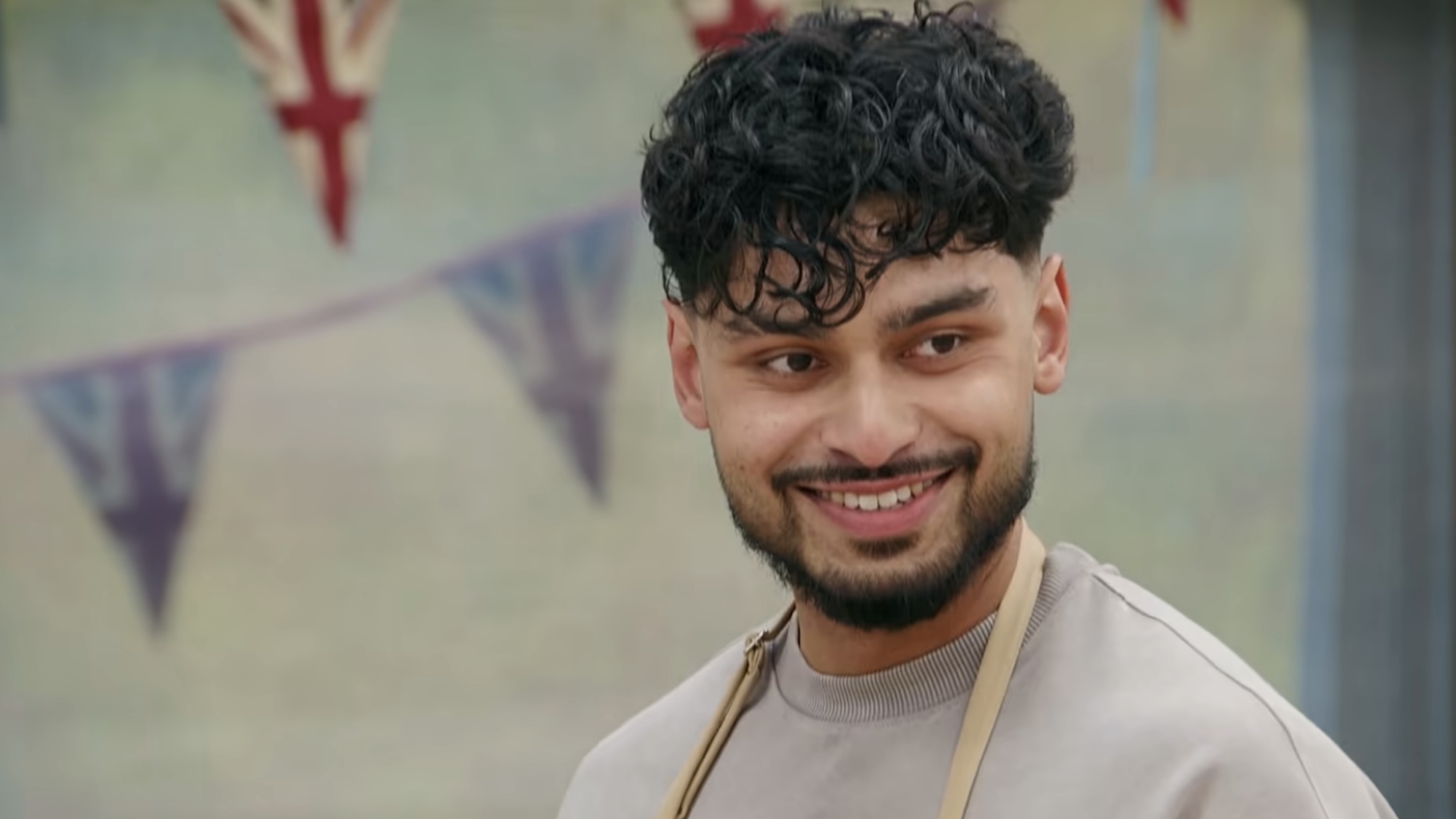 Mo Mahirr smiling in a grey shirt in the trailer for The Great British Bake Off.