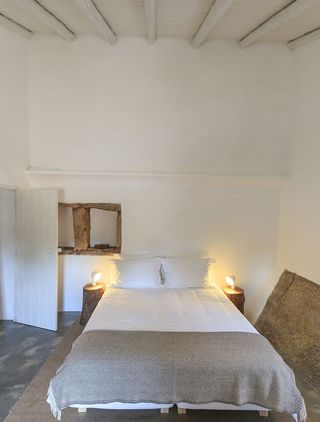 Bedroom with bed with white linen next to two tree stumps with lamps on it and a white door