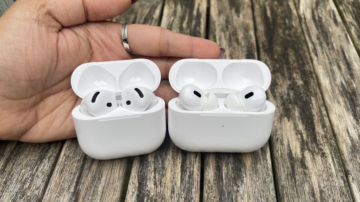 The AirPods 4 and AirPods Pro 2 in their charging cases standing side by side on a wooden surface with a hand out flat behind them.