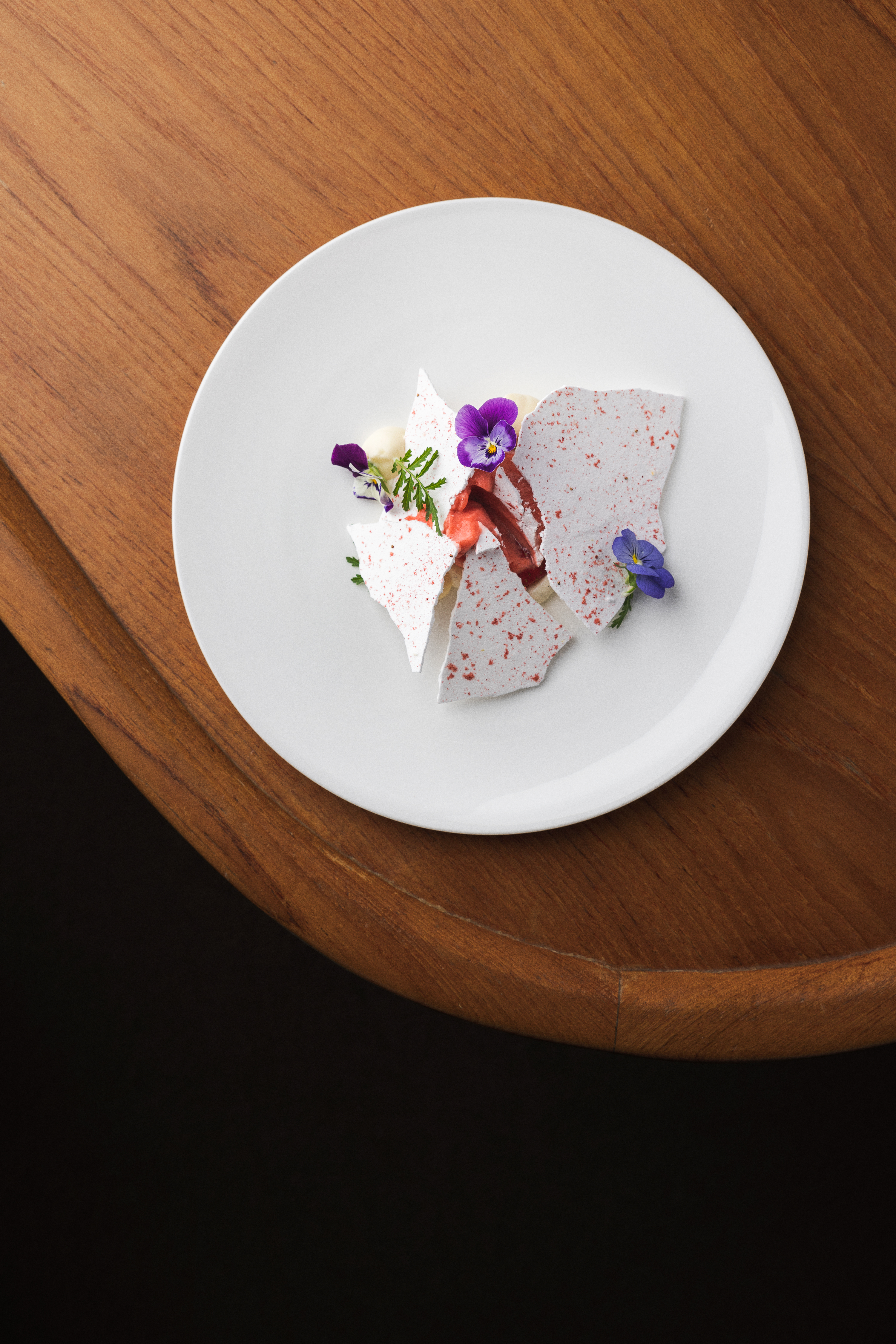 A soft-edged wooden table topped by a round, white ceramic plate where a deconstructed dessert is decorated with fresh herbs and purple flowers.