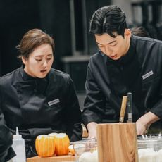 chefs in black coats preparing food over a silver counter top in an open kitchen in a still from the netflix korean reality show culinary class wars season 2