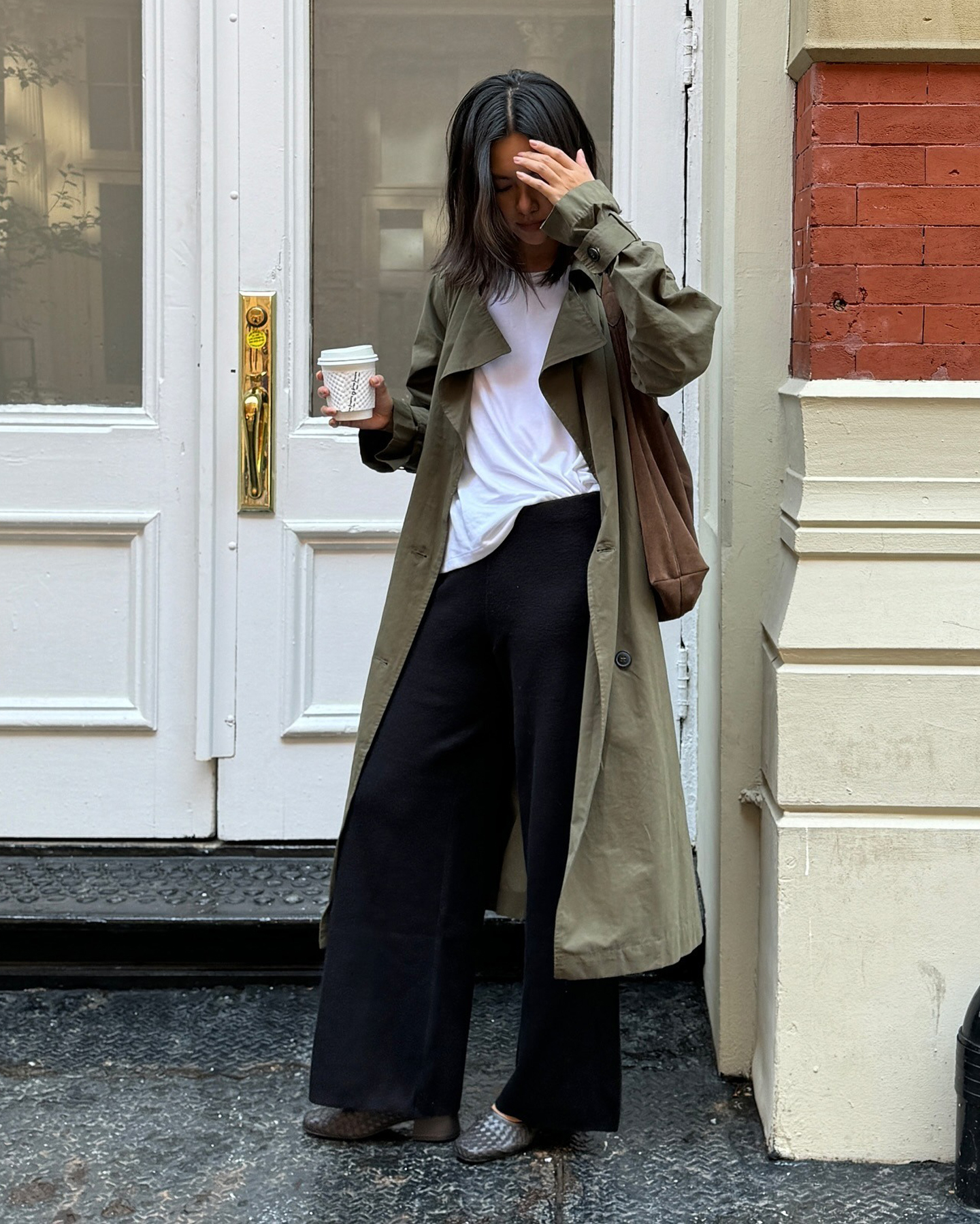 NYC style influencer Mimi Nguyen poses on a street in Manhattan wearing a green trench coat, brown suede tote bag, black knit wide-leg pants, and gray woven heels
