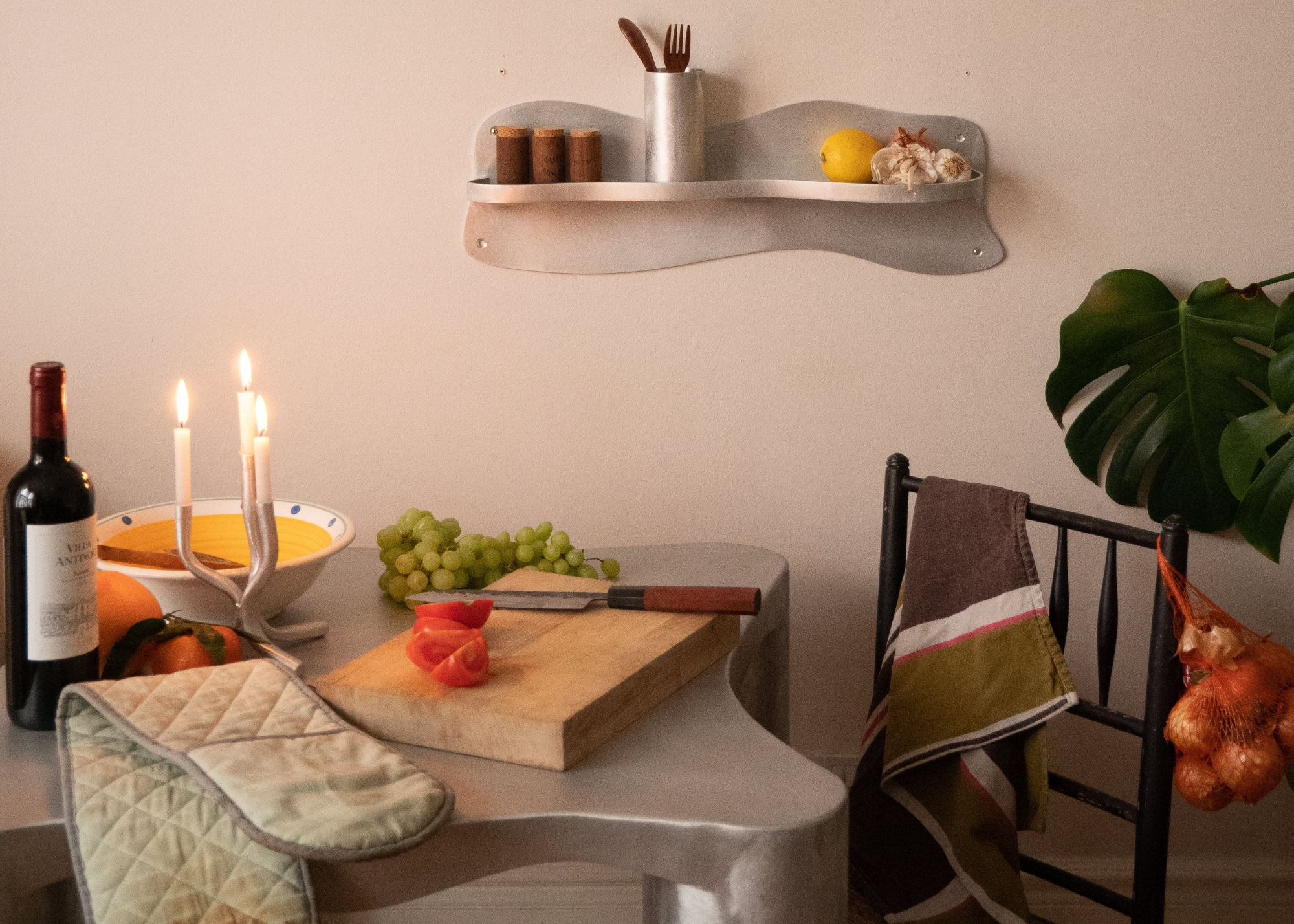 A brushed metal table with a wooden cutting board, grapes, halved tomatoes, oven mitts, a candle holder, a bottle of wine, and a floating shelf on the wall