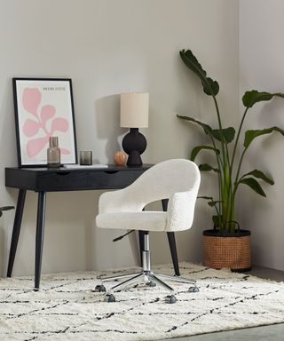 A home office with greige walls with a black desk with a lamp, vases, and pink leaf wall art on it, a white boucle office chair next to it and a tall leafy plant in a rattan basket to the right of it, and a white rug