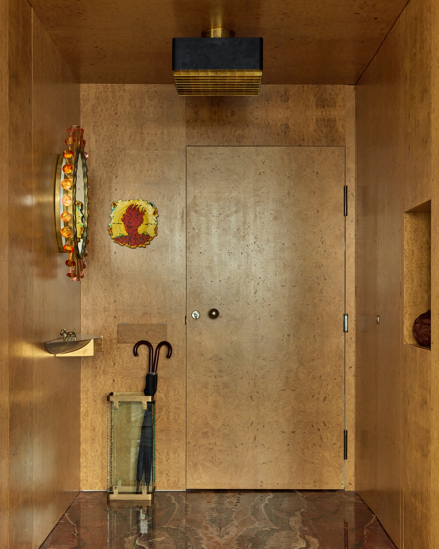 Image of a luxurious entryway drenched in large wooden paneling. There is a glass umbrella holder and an ornate orange mirror on the wall.