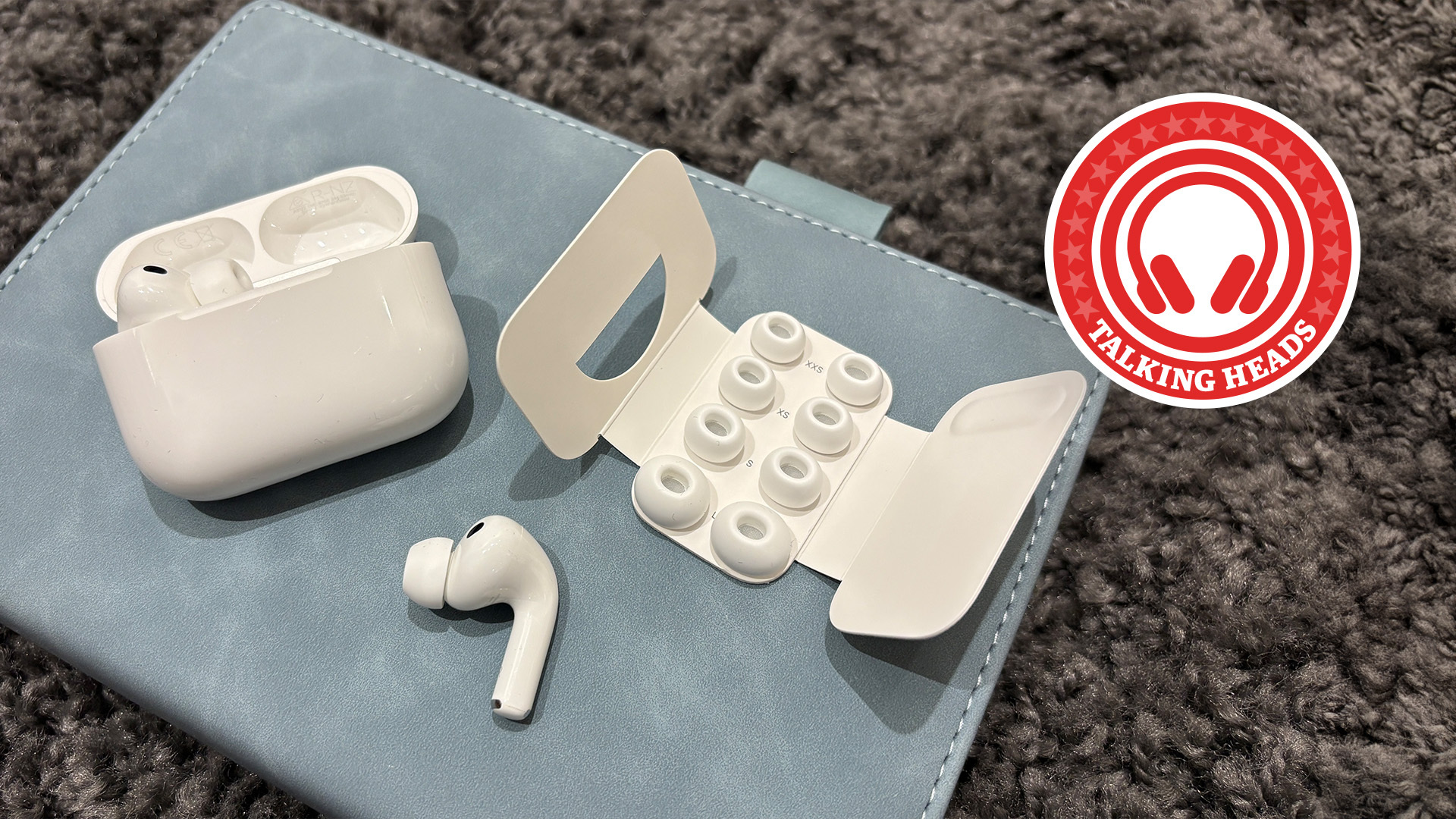 AirPods Pro 3 case, tips and earbuds on a blue notebook with a grey carpet in the background