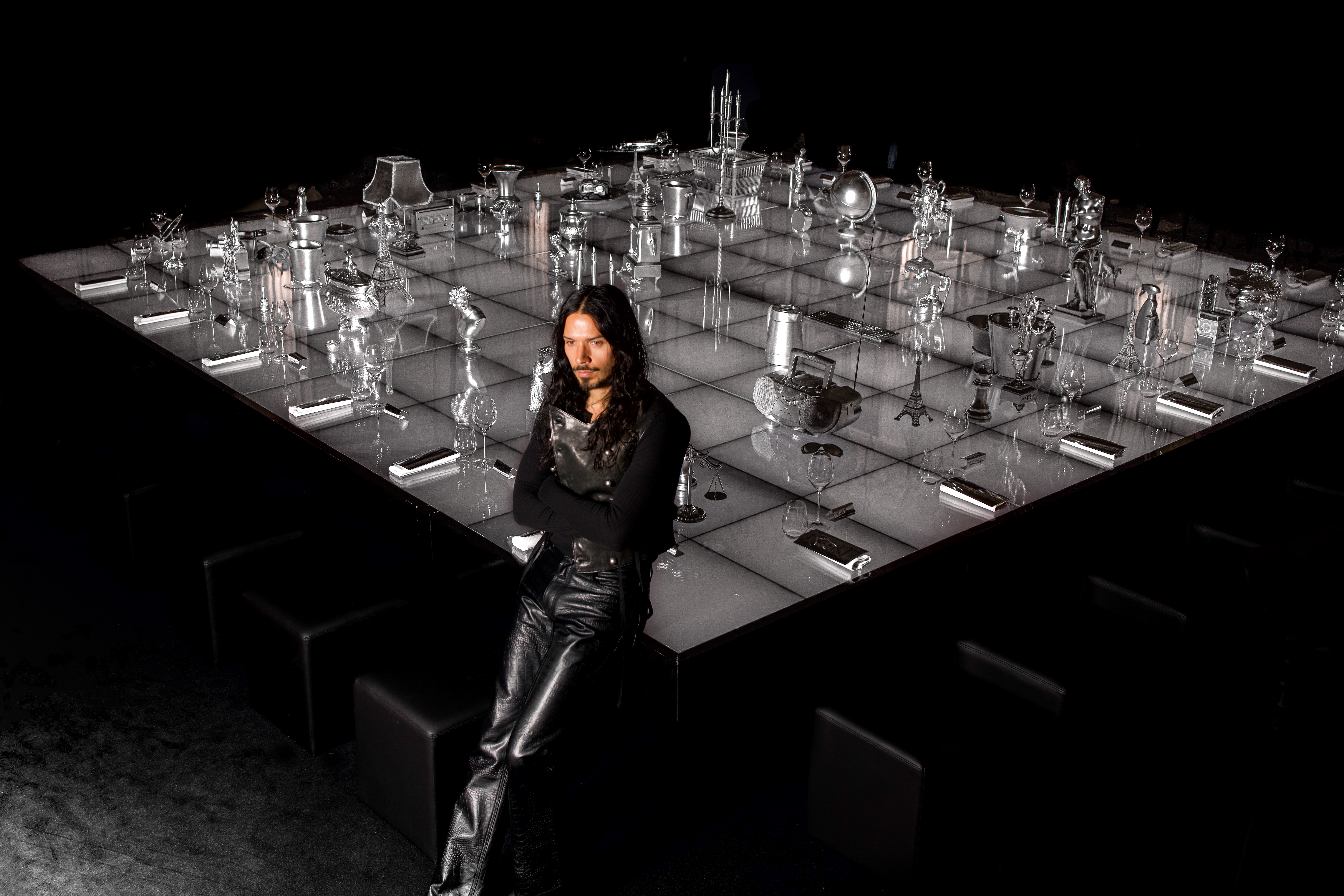 A monochromatic installation set inside a palatial hotel with mural ceilings, featuring a back-lit white surface topped by lots of silver objects, around which a man with dark long hair poses.