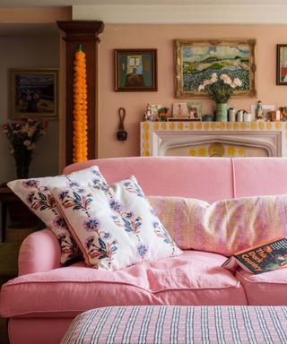 A blush pink couch with two white floral throw pillows on it, with a mantel behind it with wall art on it and above it on a peach wall, and a vase of roses on top of it