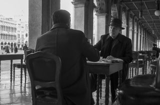 a man (John Malkovich as reeves) sits with another man at an outdoor cafe in venice, in 'ripley'