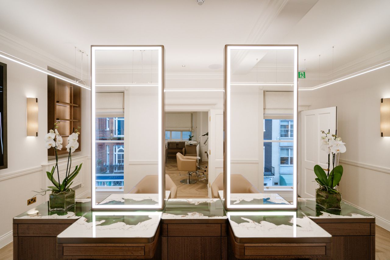 A brightly lit hair salon features beautifully crafted hairdressing stations in dark wood and glass, with abstract plaster and marble countertops, light-integrated mirrors, orchids, and cream side sconces.