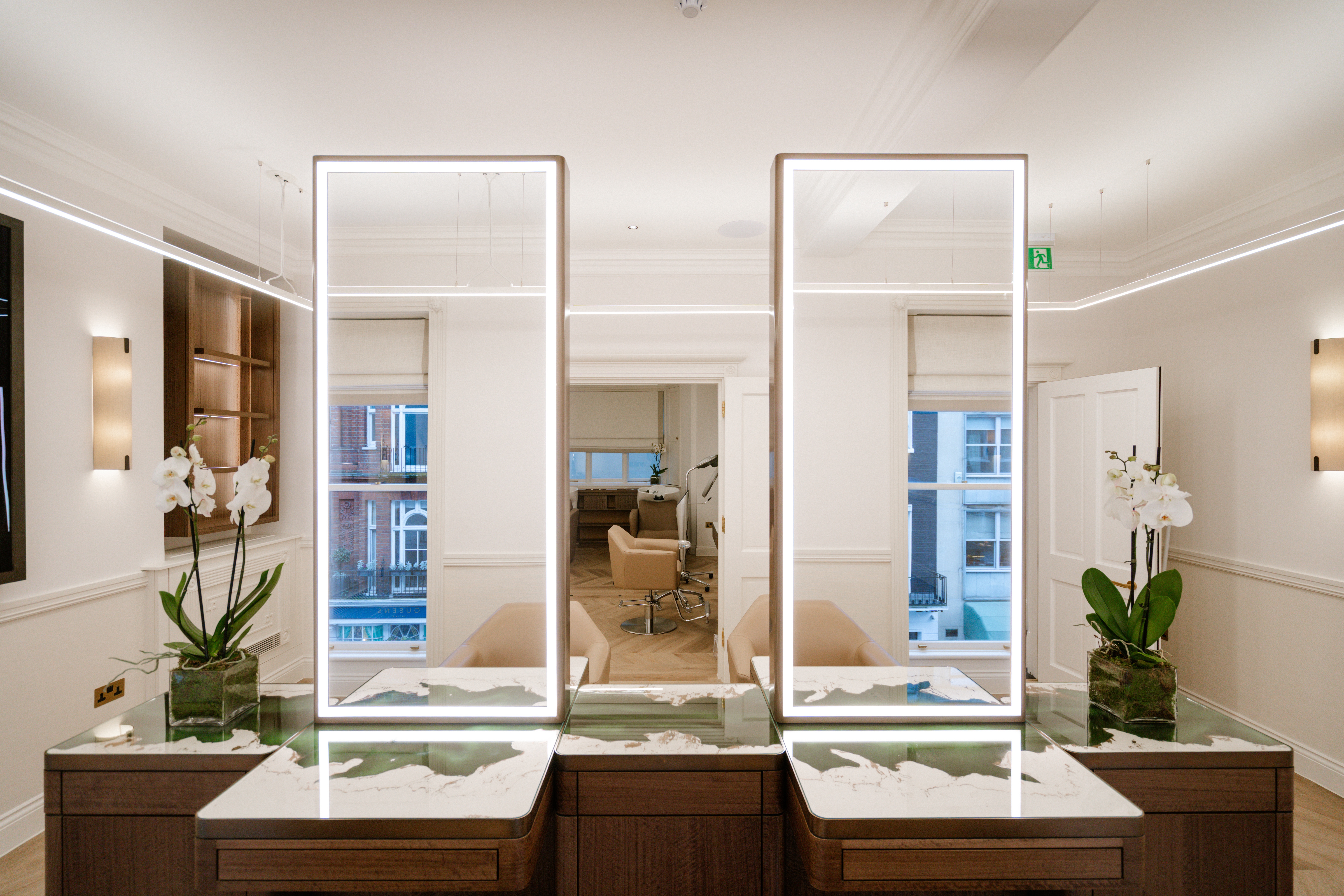 A brightly lit hair salon features beautifully crafted hairdressing stations in dark wood and glass, with abstract plaster and marble countertops, light-integrated mirrors, orchids, and cream side sconces.