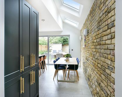 An example of side return extension ideas showing a kitchen extension with dark green cabinets and exposed brickwork behind a wooden dining table with blue chairs