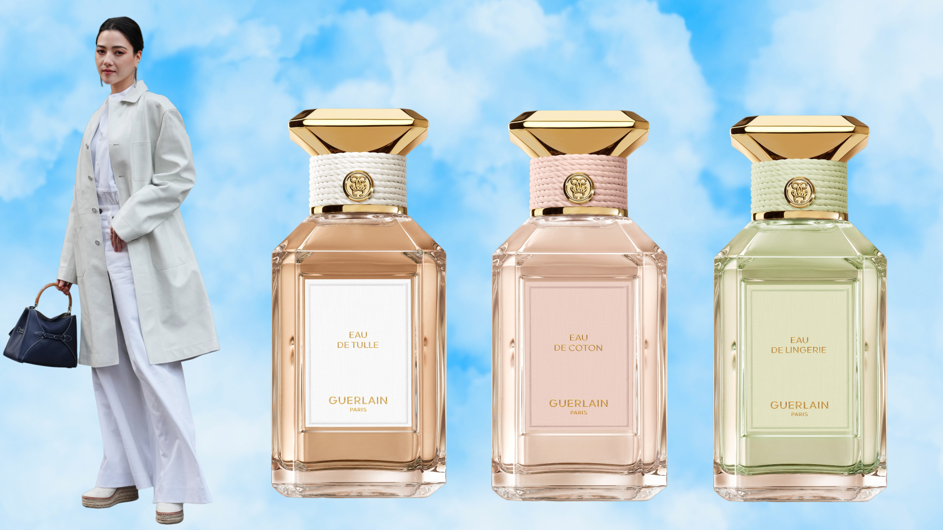 A woman wearing a white jacket and pants next to three bottles of Guerlain perfume. A blue sky with clouds backdrop is behind them all. 