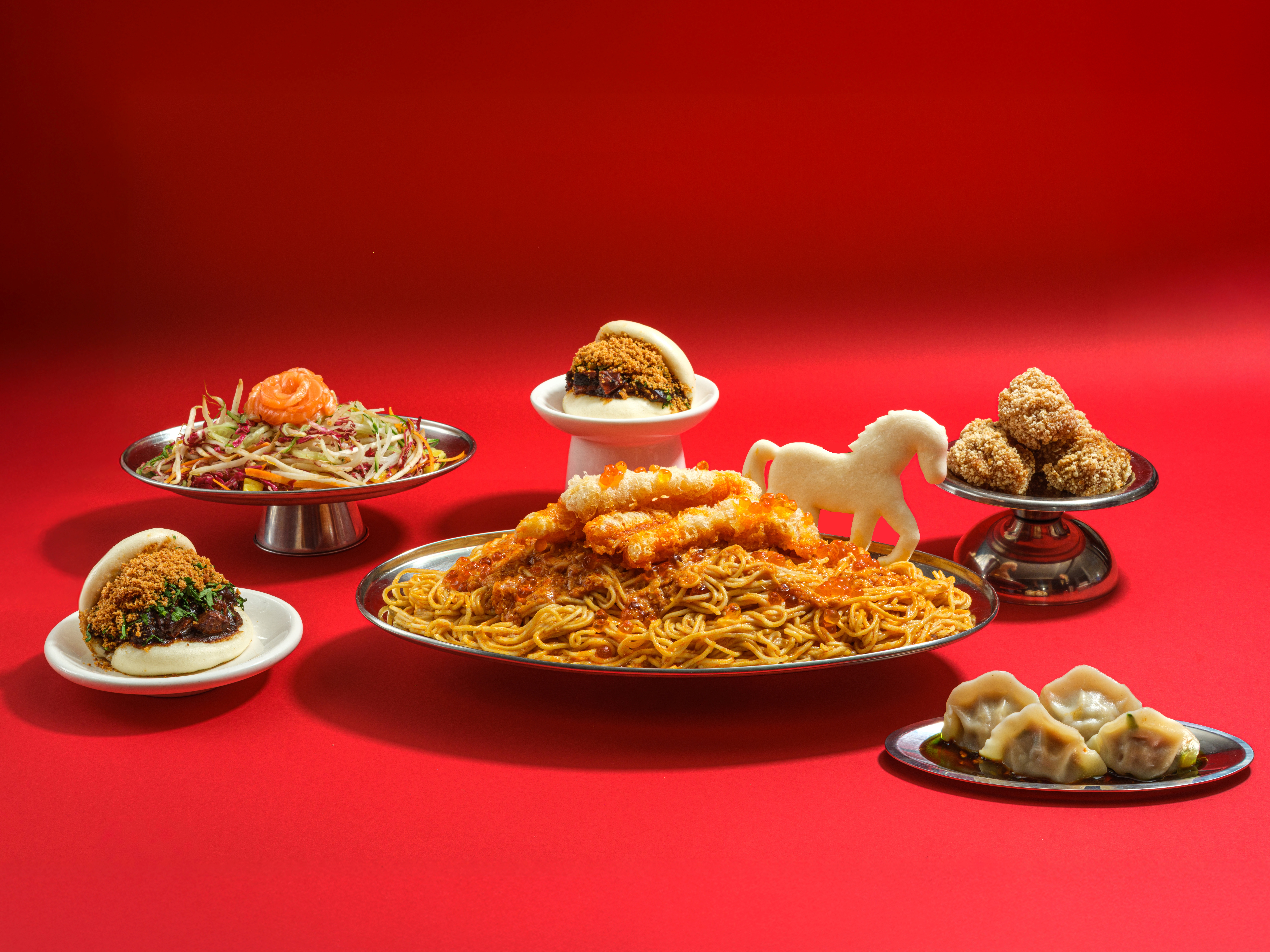 A series of beautifully plated food plates, including noodles, dumplings, baos, and fried croquettes, sit in silver plates against a red background, the largest, central of them decorated with horse-shaped dough.