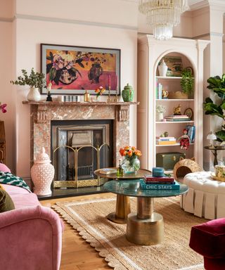 A very light pink living room with a brown marbled fireplace, bookshelf, coffee table, and chairs