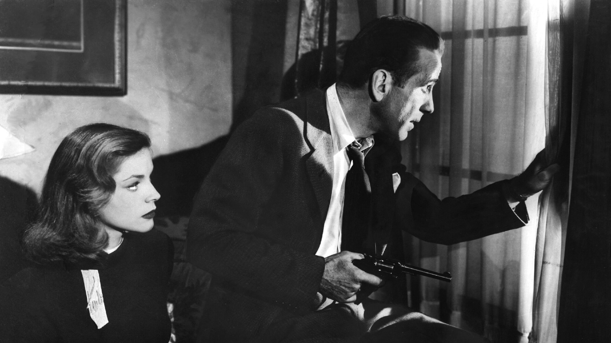 a still from the movie 'The Big Sleep.' it is in black and white, with Lauren Bacall at left, seated, and Humphrey Bogart looking out a window with a gun in his hand