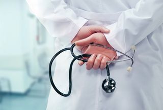 A doctor stands holding a stethoscope behind his back.