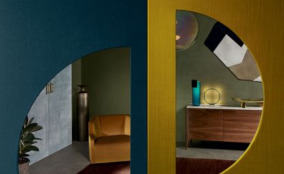 Interior living space, two wood doors, one half blue the other yellow, half circle glass with view inside room, brown arnchair, wooden cabinet, lighting, pot plants, grey floor, brwon rug, glass ball ceiling light, wall art, pale green walls
