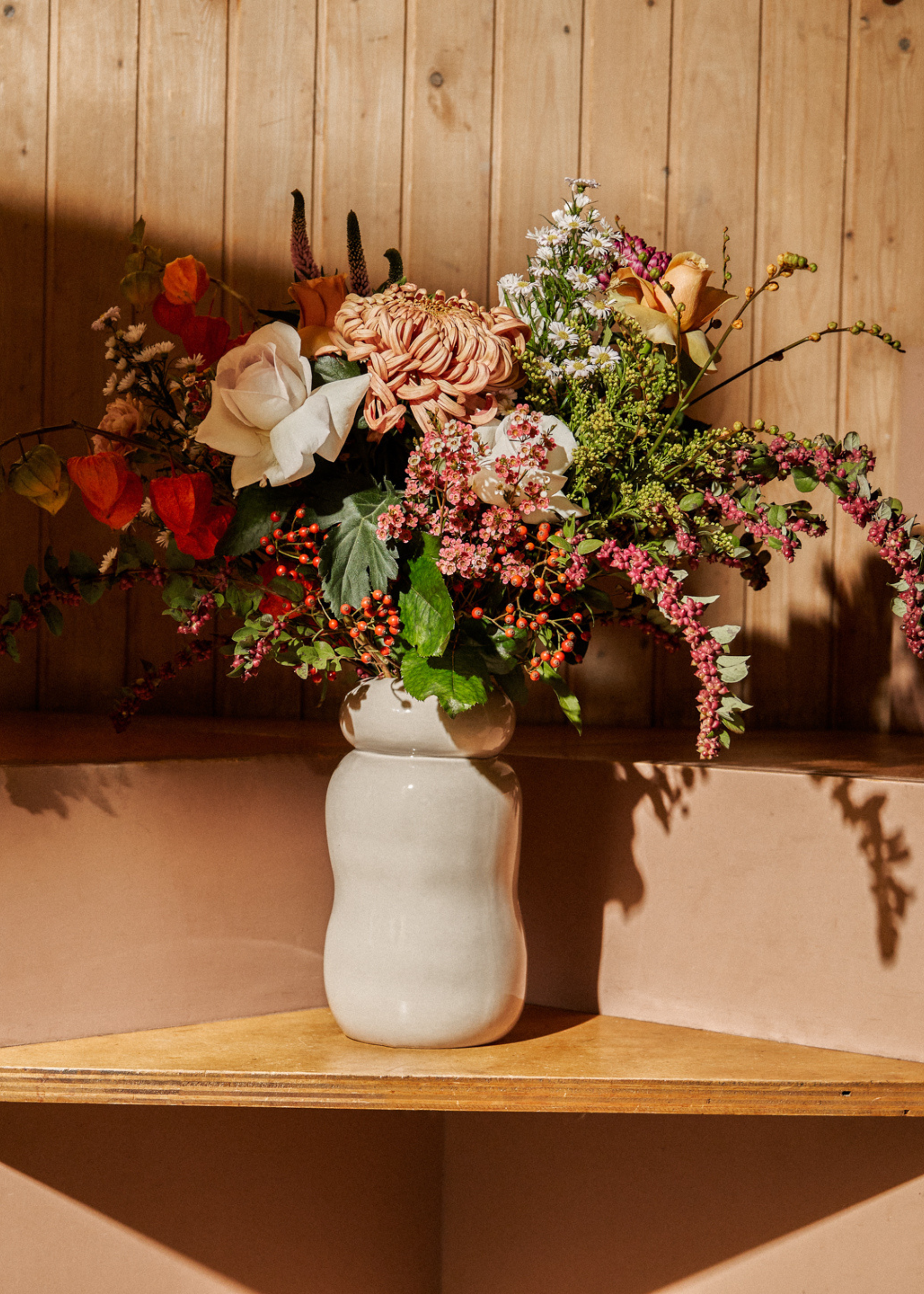 A ceramic vase with aster flowers and other textured flowers on a wood shelf
