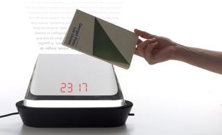A digital bedside clock , with a hand holding a book over it.