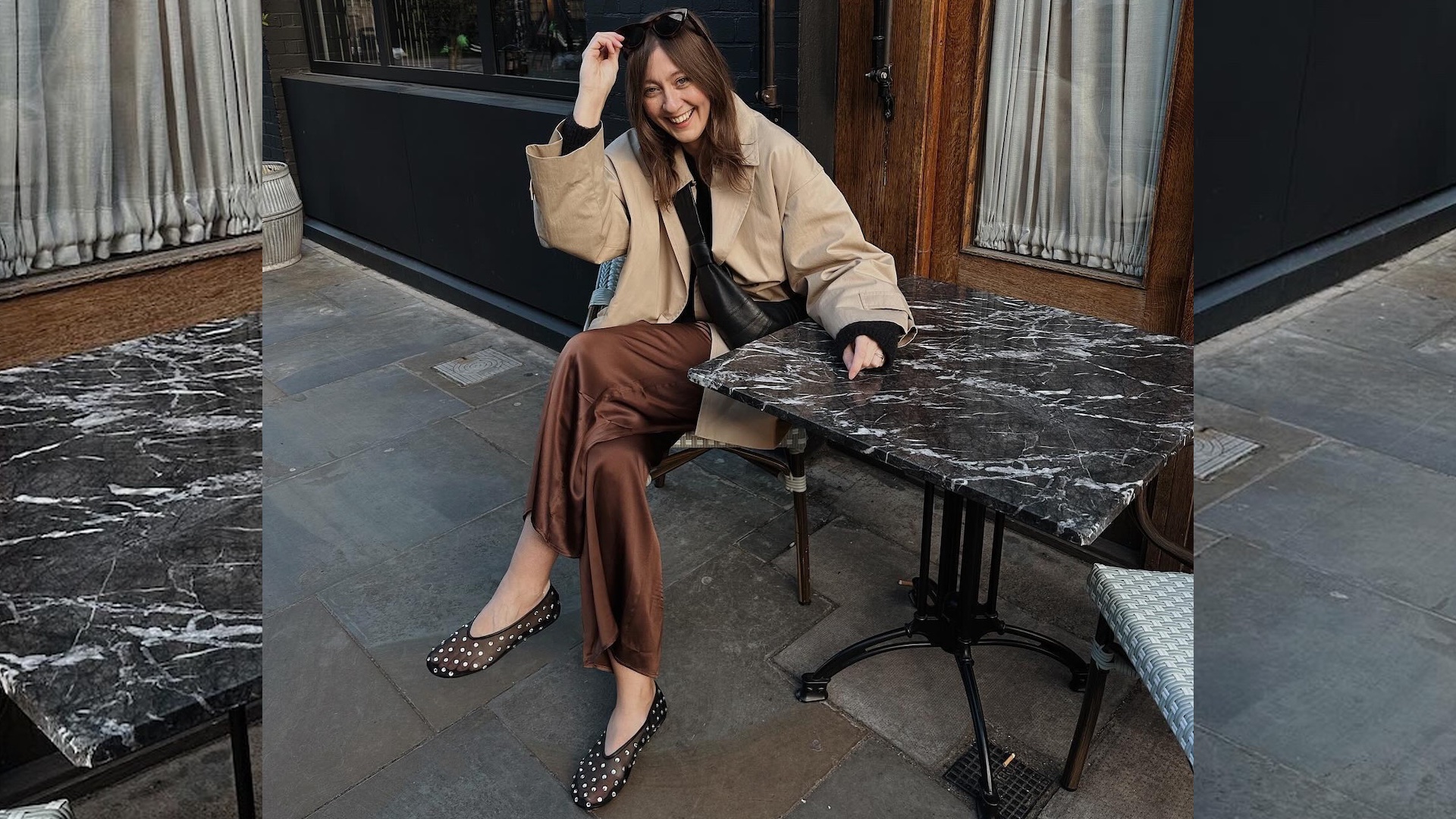 British style influencer Rachael Keegan sits at an outdoor marble cafe table wearing a cropped trench coat, brown satin pants, and studded black ballet flat