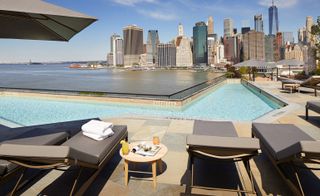 View of an outdoor area at 1 Hotel Brooklyn Bridge, New York, US featuring a pool, grey lounge chairs, a round table and grey parasols. There is also a view of the East River and tall buildings that are nearby