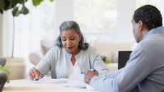 An older woman looks at paperwork with a financial adviser at her dining room table.