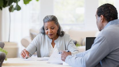 An older woman looks at paperwork with a financial adviser at her dining room table.