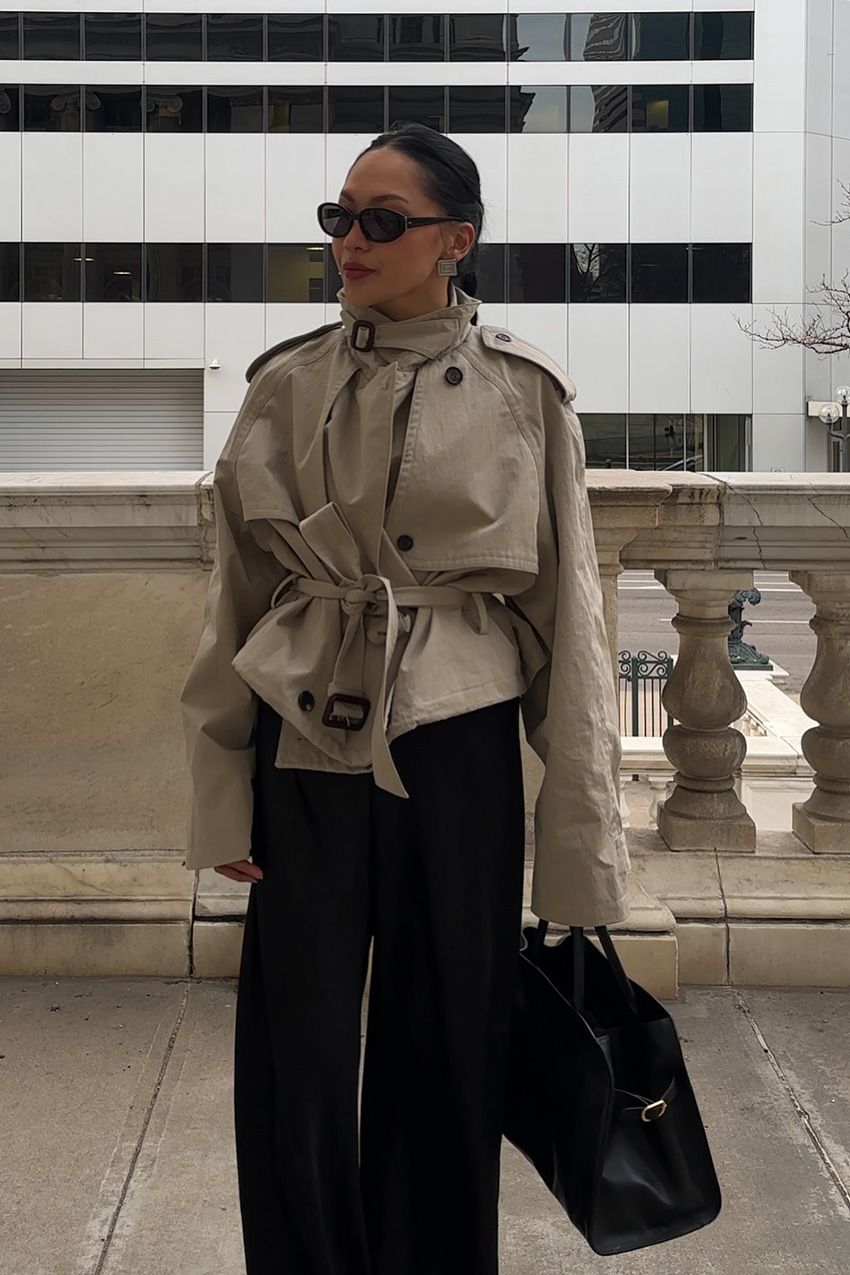 A woman standing outside wearing a tan cropped funnel neck trench coat styled with black wide-leg pants, a black bowler bag, silver square earrings, and black oval sunglasses.