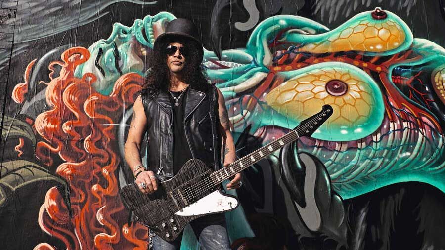 Slash standing against a grafitti&#039;d wall with a guitar