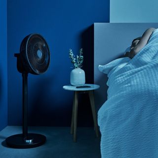 Duux standing fan in blue bedroom at night while person sleeps in bed closeby