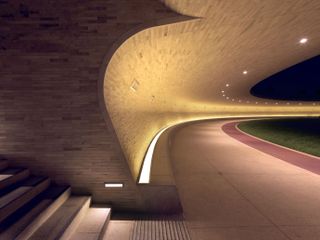 Night time image of oxygen park Qatar's walkway, brick walls, curved design with spot lighting, stone steps to the left, guiding light around the edge of the wall, neutral stone slab path with red stripe and grass verge to the right