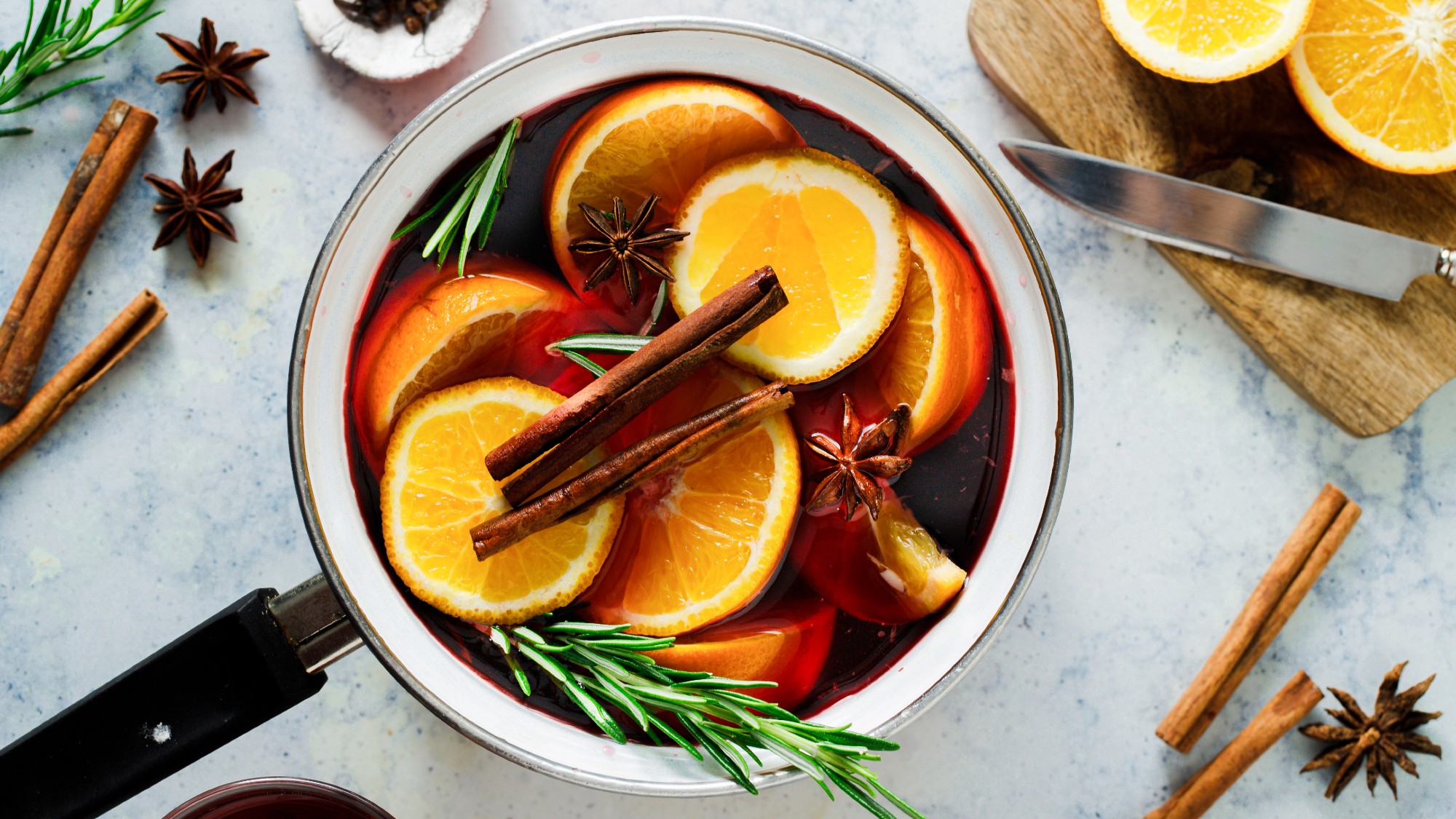 overhead shot of a saucepan filled with red wine cinnamon sticks, rosemary sprigs and lots of sliced orange