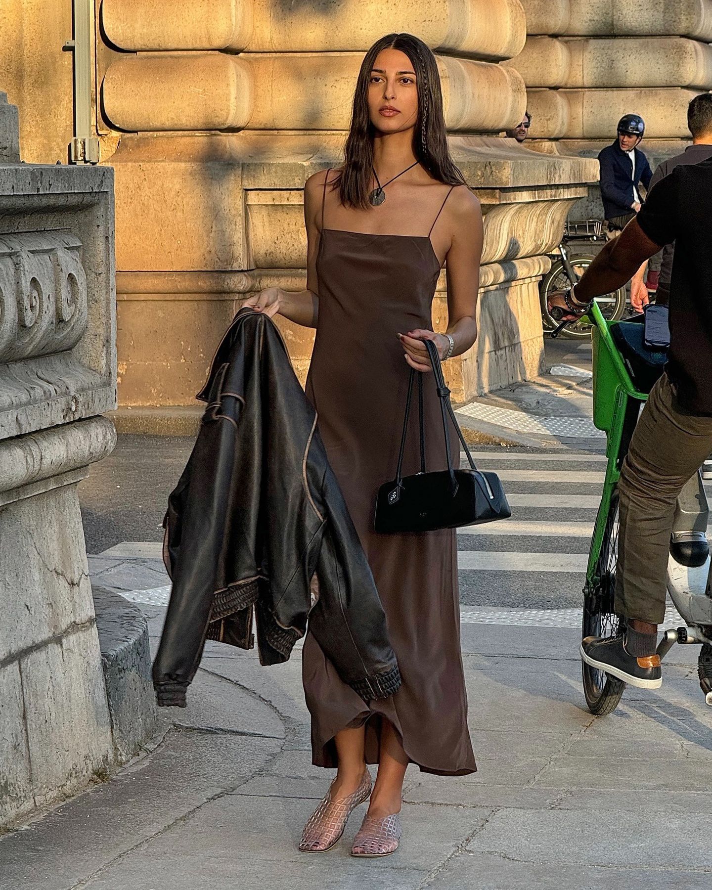 French fashion influencer Salomé Mory walking on a Paris sidewalk at sunset wearing a black cord necklace with a disc pendant, brown slip dress, black Alaia east west shoulder bag, a brown leather jacket in her hand, and clear jelly flats