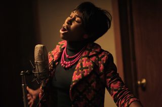 Cynthia Erivo as Aretha Franklin in the 1960s, wearing a multicoloured jacket, a black turtleneck and three strings of red beads, standing in a recording studio singing into the microphone with her head tilted back and her eyes closed