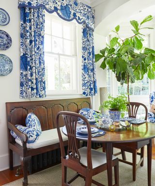 A breakfast room with wooden dining bench, table and chairs, with blue and white floral fabric on armchair, curtains and cushions
