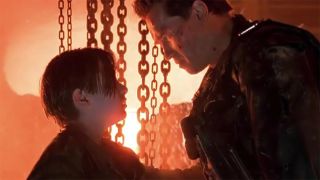 Still from the movie "Terminator 2: Judgment Day." The Terminator (a robot who looks like a muscly human male wearing a leather jacket) has his hand on a young boy's shoulder as he looks directly into his face. In the background the there are dangling thick chains and molten metal being poured.