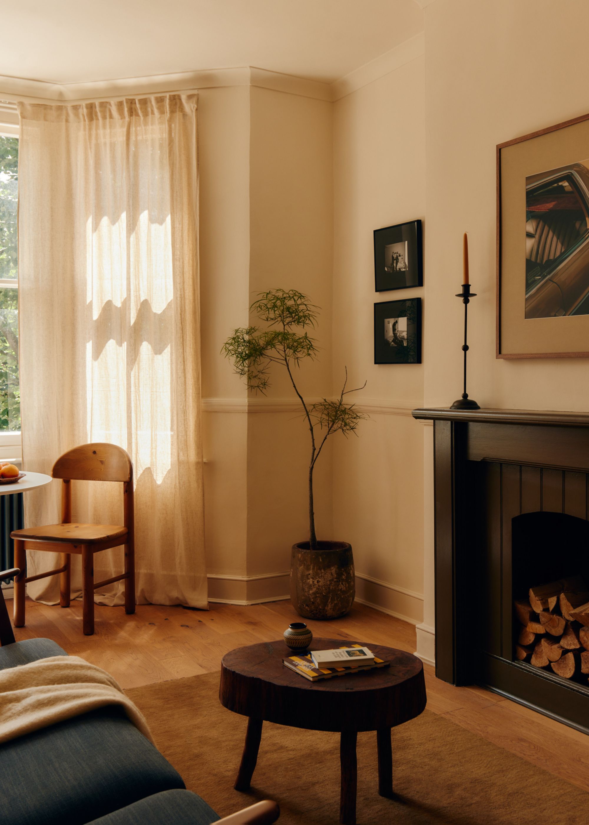 A neutral living room color-drenched an off-white with a bay window with sheer curtains, a dark fireplace, wood flooring and a dark wood coffee table.