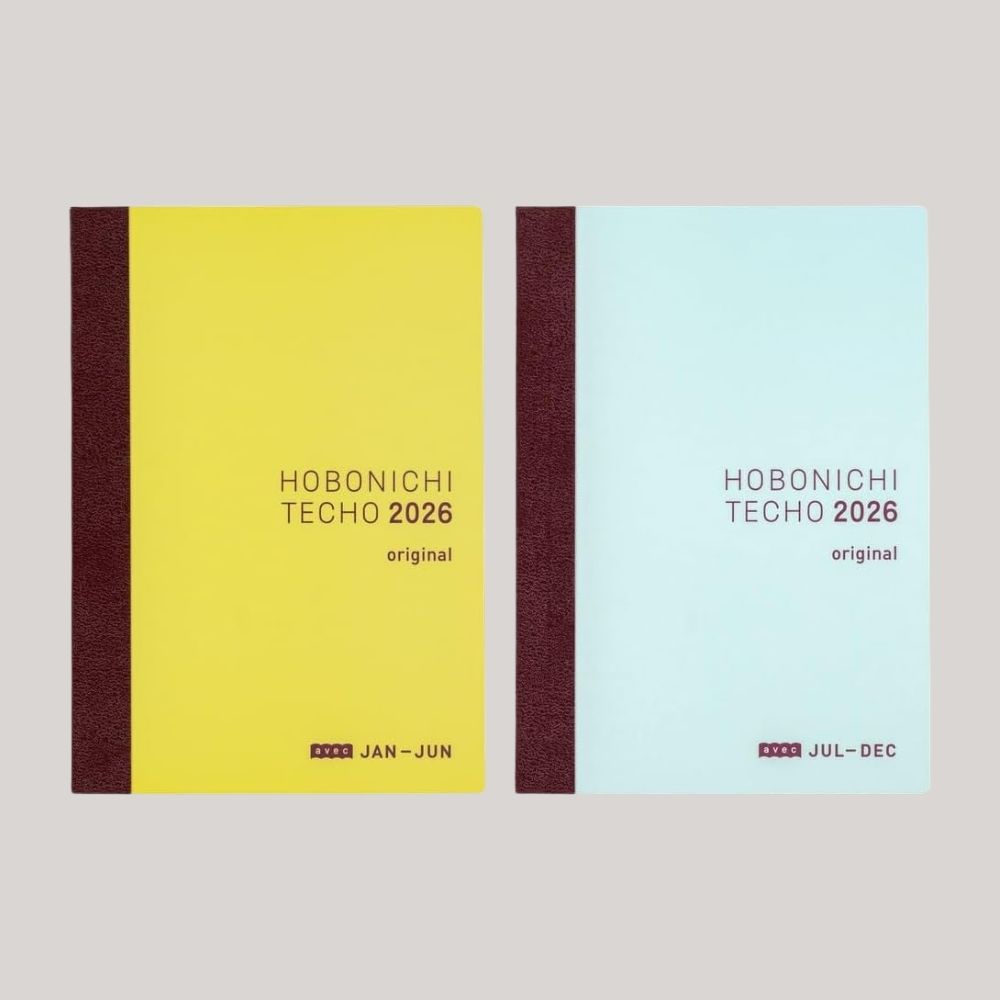 Two identical notebooks, one cherry red and yellow, the other cherry red and pale blue, read Hobonichi Techo 2026 on their cover while sitting against a pale grey background.