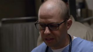 Jon Cryer with a bald head, wearing glasses and looking down in NCIS