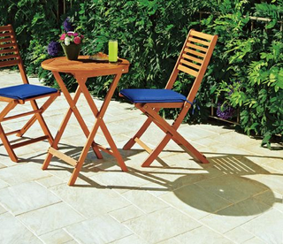 Paved patio with table and 2 chairs with blue cushions and a pot of flowers and a glass of cooldrink on the table