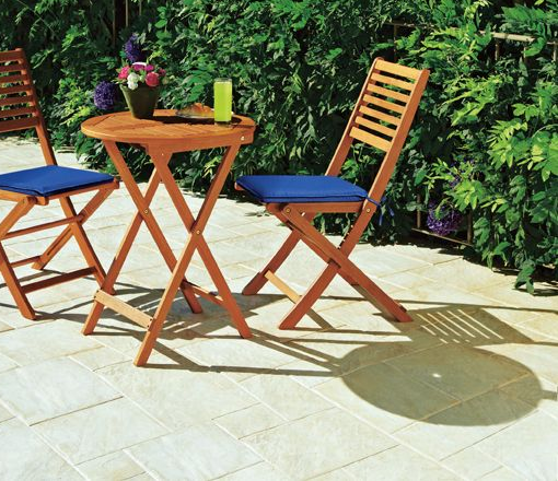 Paved patio with table and 2 chairs with blue cushions and a pot of flowers and a glass of cooldrink on the table
