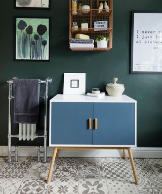 A dark green bathroom with cabinet shelves, wall art, and a blue and white table with decor on it