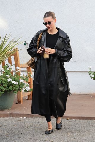 Hailey Bieber leaving her Pilates studio wearing a leather trench coat with ballet flats by The Row and a leopard Saint Laurent tote bag