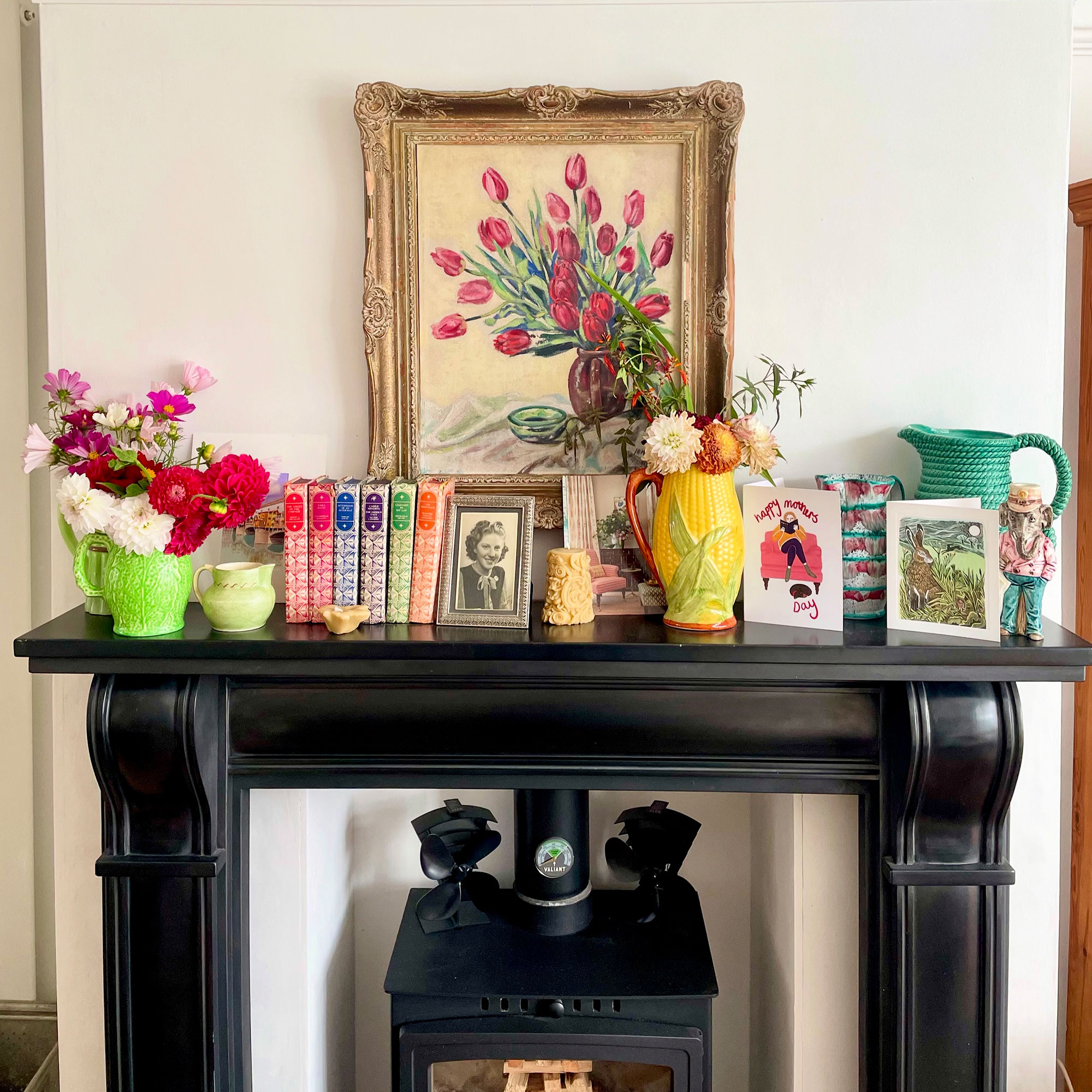 Fireplace mantlepiece with colourful books, vases filled with flowers and floral painting