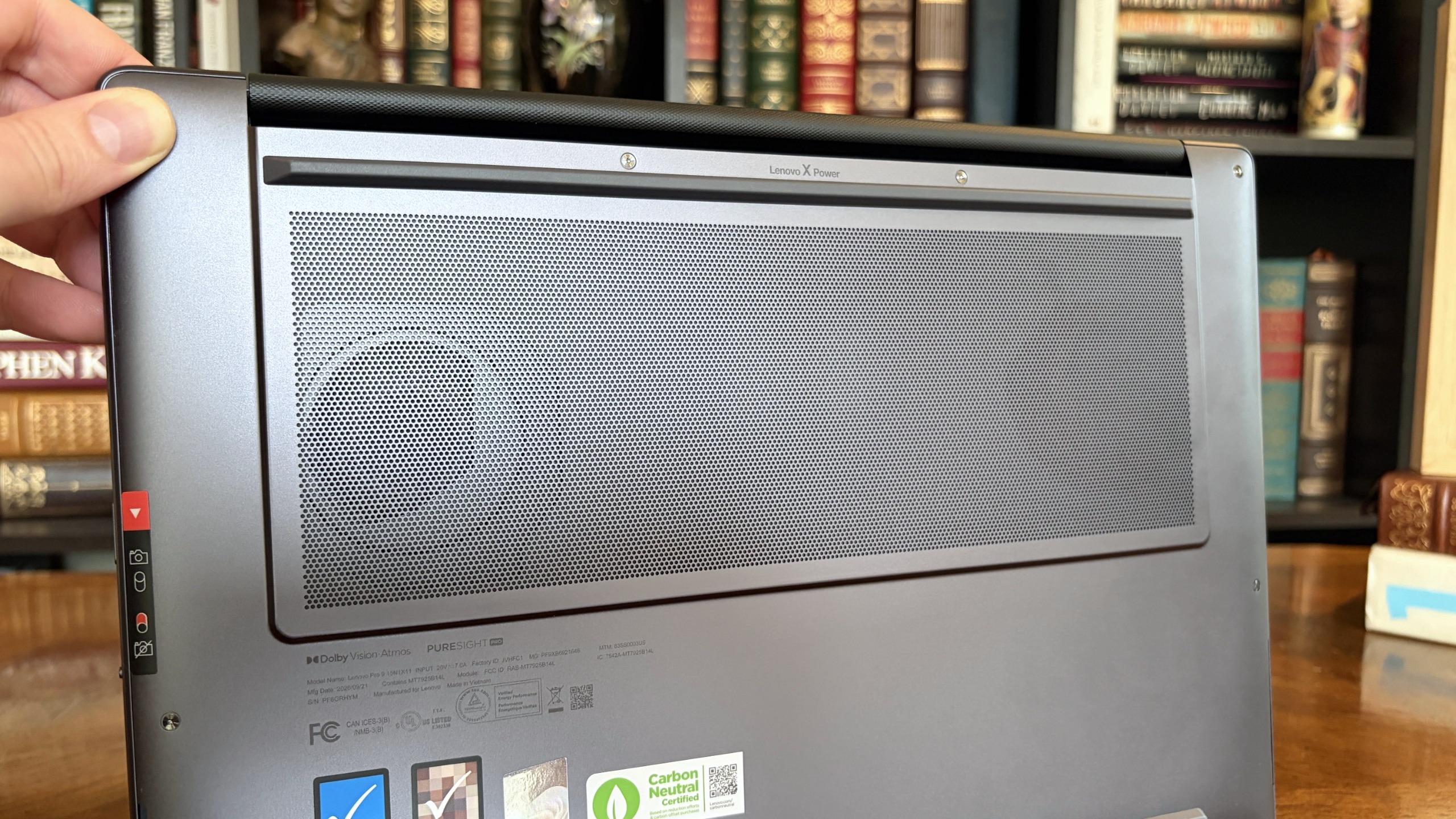 A hand holds a Lenovo Pro 9n laptop with a large mesh grille and small labels, displayed before shelves of old books in a cozy library setting.