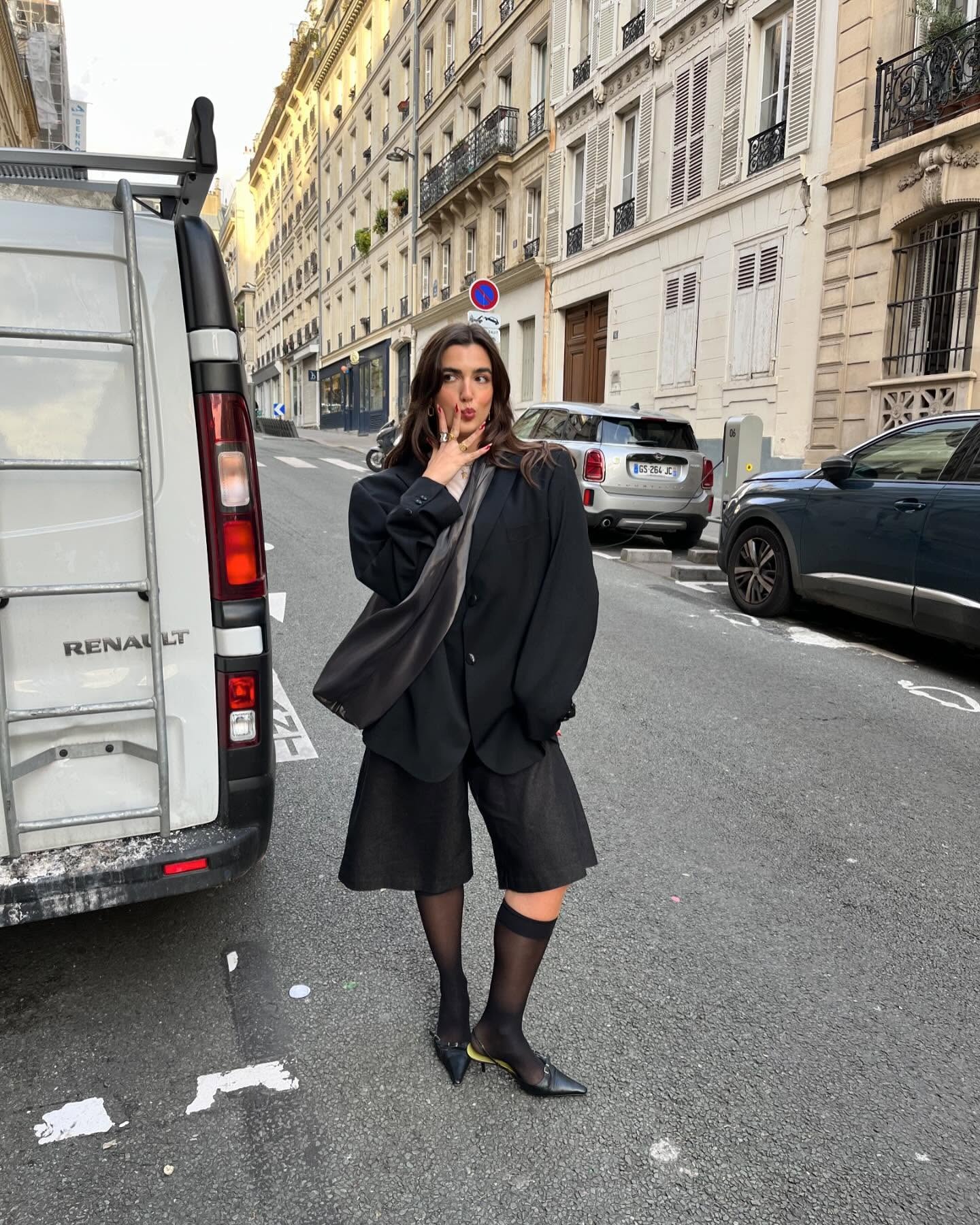Fashion person Pauline Leroy wears black mesh knee-high socks. She pairs them with black bermuda shorts and a black blazer in a recent image from her Instagram.
