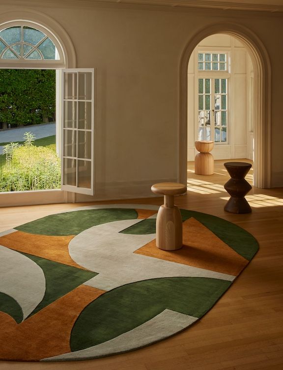 a green, cream, and yellow mosaic-like oval rug sits on a wooden floor in a room filled with wooden sculptures.